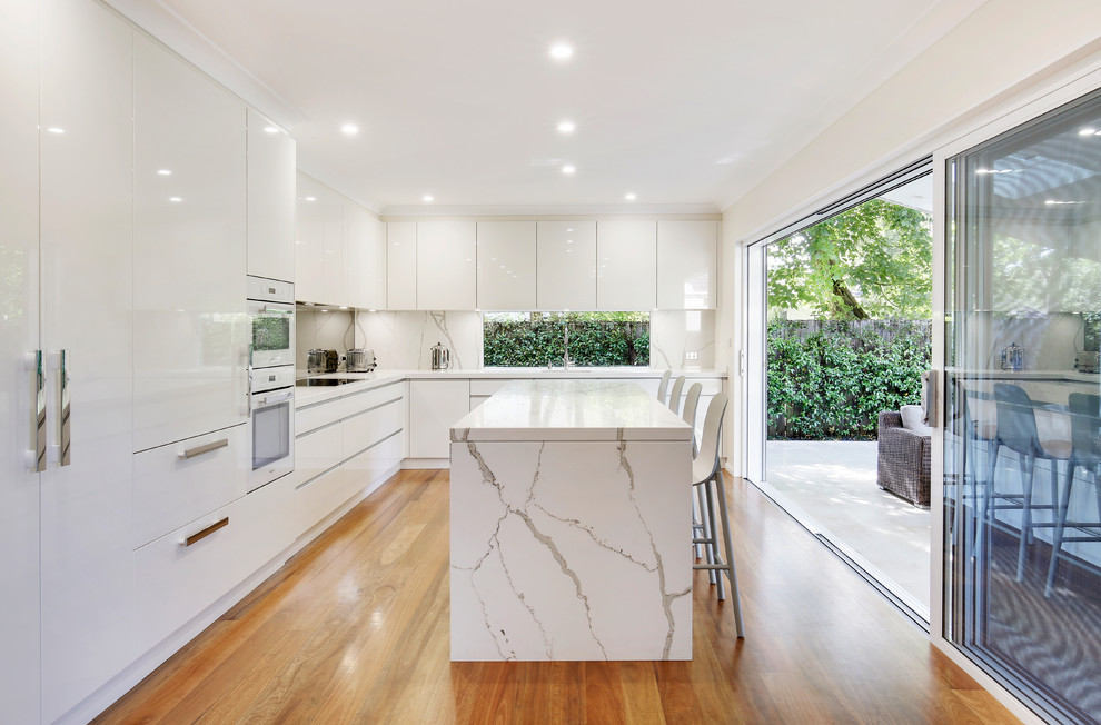This is an example of a contemporary l-shaped eat-in kitchen in Sydney with an undermount sink, flat-panel cabinets, white cabinets, quartzite benchtops, white splashback, white appliances, medium hardwood floors, with island, brown floor and white benchtop.