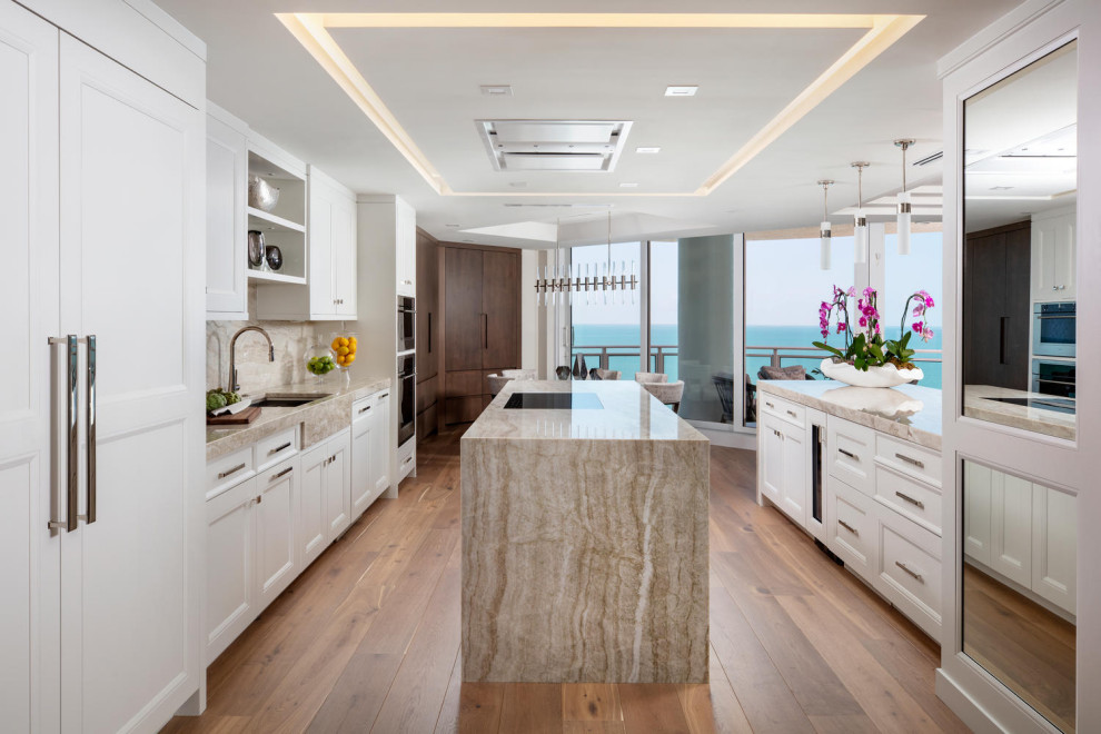 Photo of a large beach style eat-in kitchen in Miami with an undermount sink, recessed-panel cabinets, white cabinets, quartzite benchtops, beige splashback, stone slab splashback, stainless steel appliances, medium hardwood floors, multiple islands, brown floor and beige benchtop.