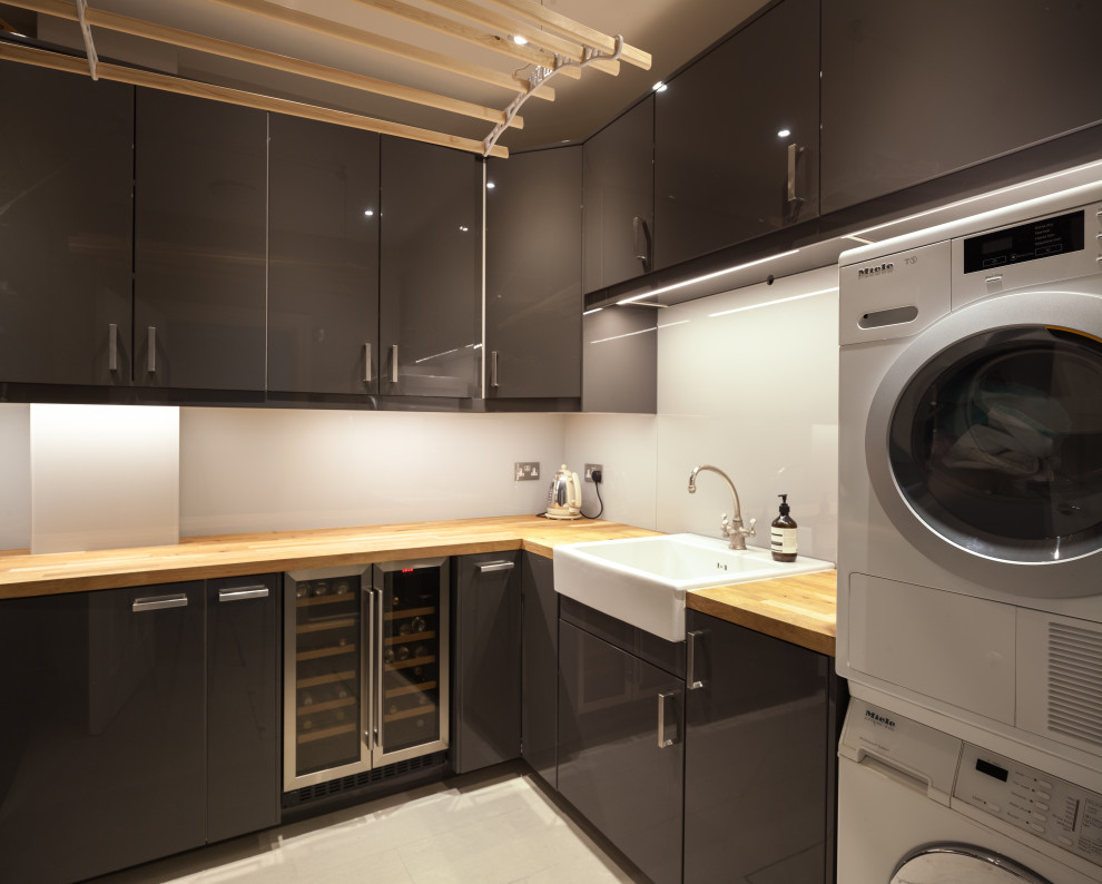 Photo of a small modern u-shaped utility room with a farmhouse sink, flat-panel cabinets, grey cabinets, wood benchtops, grey splashback, glass sheet splashback, an integrated washer and dryer and beige benchtop.