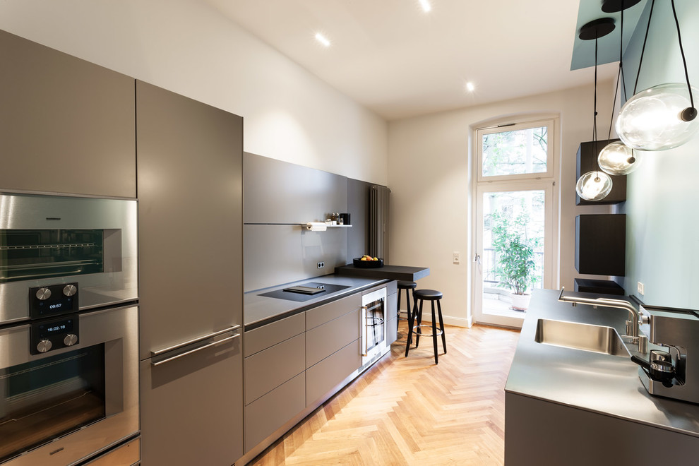 Photo of a mid-sized contemporary galley separate kitchen in Dusseldorf with an integrated sink, flat-panel cabinets, grey cabinets, grey splashback, stainless steel appliances, medium hardwood floors, no island, brown floor and grey benchtop.