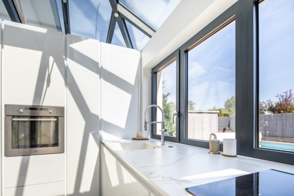 На фото: угловая кухня-гостиная среднего размера в стиле модернизм с монолитной мойкой, белыми фасадами, белой столешницей и мойкой у окна с