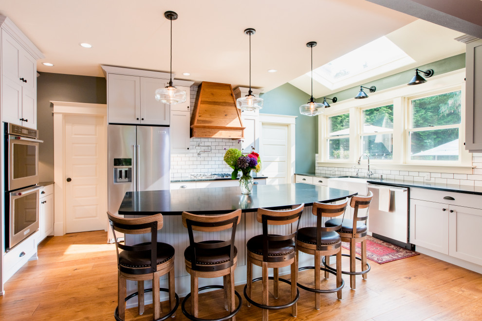Large arts and crafts u-shaped eat-in kitchen in Seattle with a farmhouse sink, shaker cabinets, white cabinets, quartz benchtops, white splashback, brick splashback, stainless steel appliances, medium hardwood floors, with island, brown floor, black benchtop and vaulted.