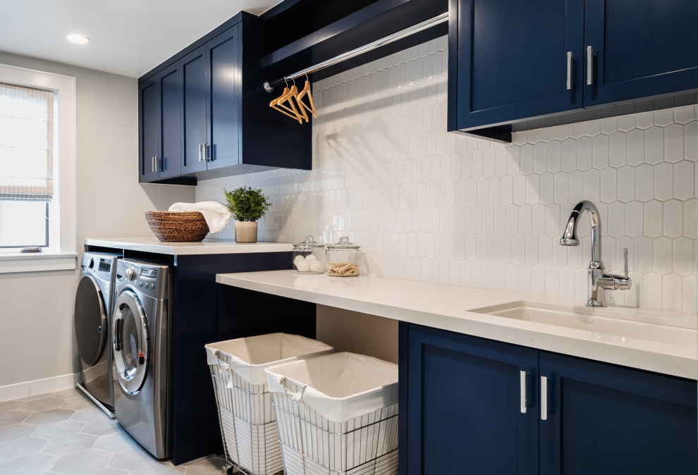 サンディエゴにある中くらいなカントリー風のおしゃれな洗濯室 (ll型、アンダーカウンターシンク、シェーカースタイル扉のキャビネット、青いキャビネット、クオーツストーンカウンター、白いキッチンパネル、セラミックタイルのキッチンパネル、グレーの壁、磁器タイルの床、左右配置の洗濯機・乾燥機、グレーの床、白いキッチンカウンター) の写真