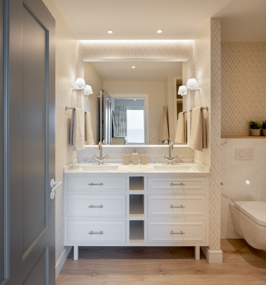 Photo of a large transitional master bathroom in Bilbao with raised-panel cabinets, white cabinets, an alcove shower, a wall-mount toilet, beige tile, porcelain tile, beige walls, laminate floors, an undermount sink, engineered quartz benchtops, brown floor, a hinged shower door, white benchtops, an enclosed toilet, a double vanity, a built-in vanity and wallpaper.