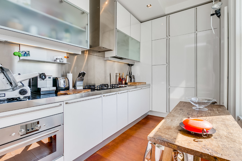 This is an example of a small eclectic l-shaped separate kitchen in London with flat-panel cabinets, white cabinets, stainless steel benchtops, metallic splashback, metal splashback, stainless steel appliances, medium hardwood floors, no island, brown floor and grey benchtop.
