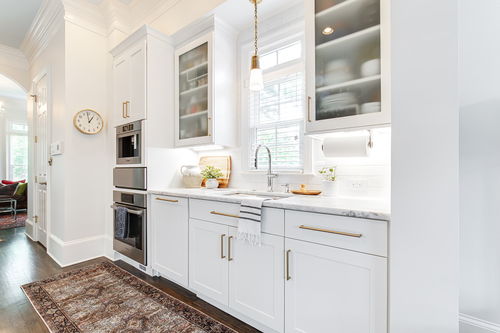 This is an example of a mid-sized eclectic galley open plan kitchen in Atlanta with an undermount sink, shaker cabinets, white cabinets, quartzite benchtops, white splashback, porcelain splashback, stainless steel appliances, medium hardwood floors, a peninsula, brown floor and white benchtop.