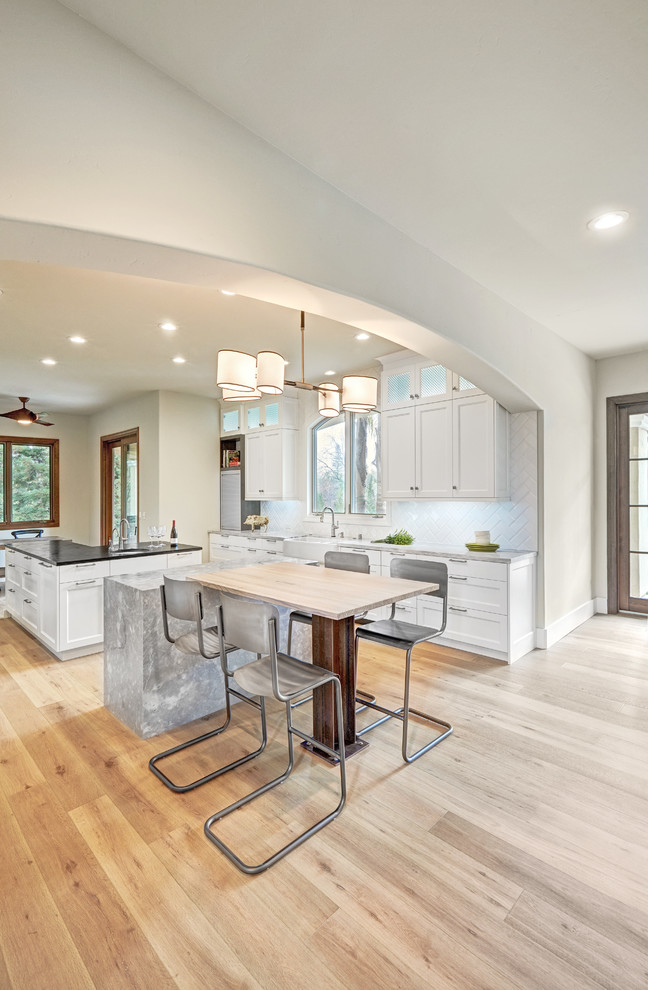 Design ideas for a large eclectic u-shaped open plan kitchen in Sacramento with a farmhouse sink, recessed-panel cabinets, white cabinets, marble benchtops, white splashback, porcelain splashback, stainless steel appliances, light hardwood floors and multiple islands.