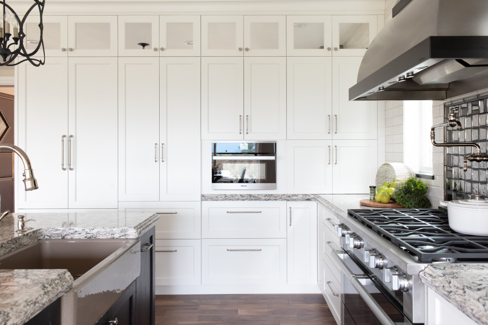 Inspiration for a large transitional l-shaped eat-in kitchen in Seattle with a farmhouse sink, white cabinets, granite benchtops, metallic splashback, glass tile splashback, stainless steel appliances, dark hardwood floors, with island, brown floor, multi-coloured benchtop and shaker cabinets.