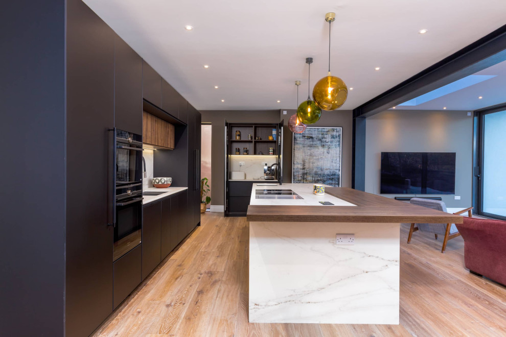 Photo of a large modern single-wall open plan kitchen in Other with flat-panel cabinets, black cabinets, quartzite benchtops, white splashback, black appliances, light hardwood floors, with island, brown floor and white benchtop.