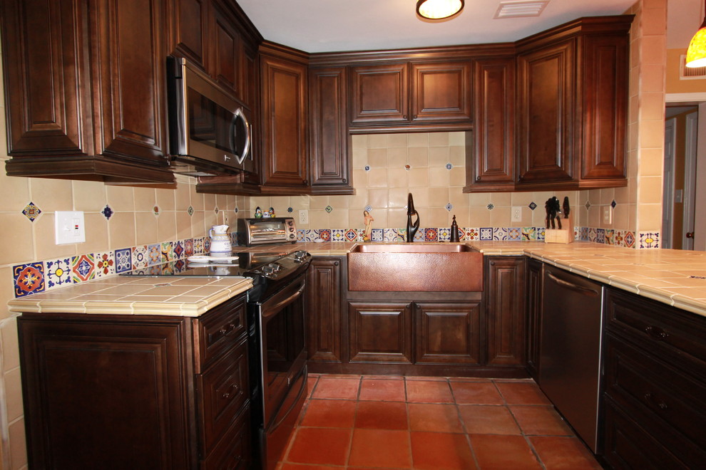 Photo of a mid-sized country u-shaped eat-in kitchen in Tampa with a farmhouse sink, raised-panel cabinets, medium wood cabinets, tile benchtops, beige splashback, ceramic splashback, stainless steel appliances and terra-cotta floors.