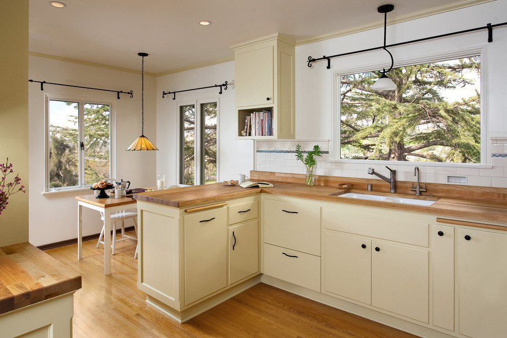 This is an example of a mid-sized traditional u-shaped eat-in kitchen in Los Angeles with an undermount sink, raised-panel cabinets, yellow cabinets, wood benchtops, white splashback, ceramic splashback, black appliances, medium hardwood floors, no island and orange floor.