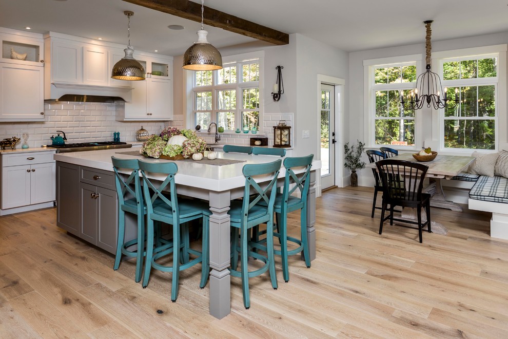 Large arts and crafts u-shaped open plan kitchen in Portland Maine with a farmhouse sink, recessed-panel cabinets, white cabinets, quartz benchtops, white splashback, subway tile splashback, stainless steel appliances, light hardwood floors and with island.