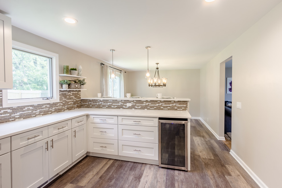This is an example of a mid-sized modern l-shaped eat-in kitchen in New York with a single-bowl sink, shaker cabinets, white cabinets, quartz benchtops, beige splashback, mosaic tile splashback, white appliances, vinyl floors, a peninsula, beige floor and white benchtop.