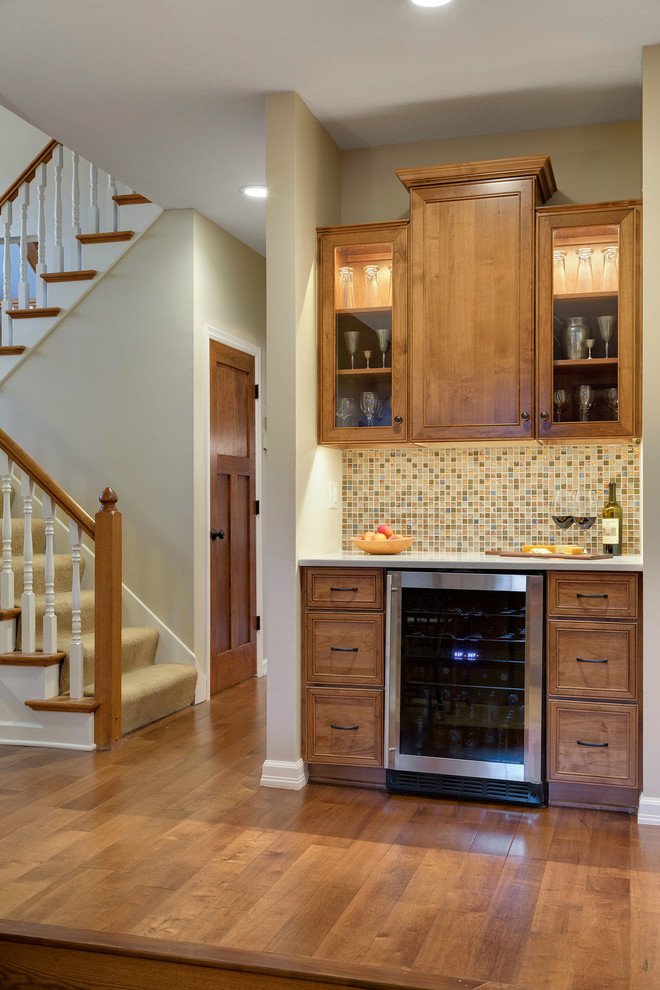 This is an example of a mid-sized traditional single-wall wet bar in Minneapolis with shaker cabinets, medium wood cabinets, solid surface benchtops, multi-coloured splashback, mosaic tile splashback, dark hardwood floors and brown floor.