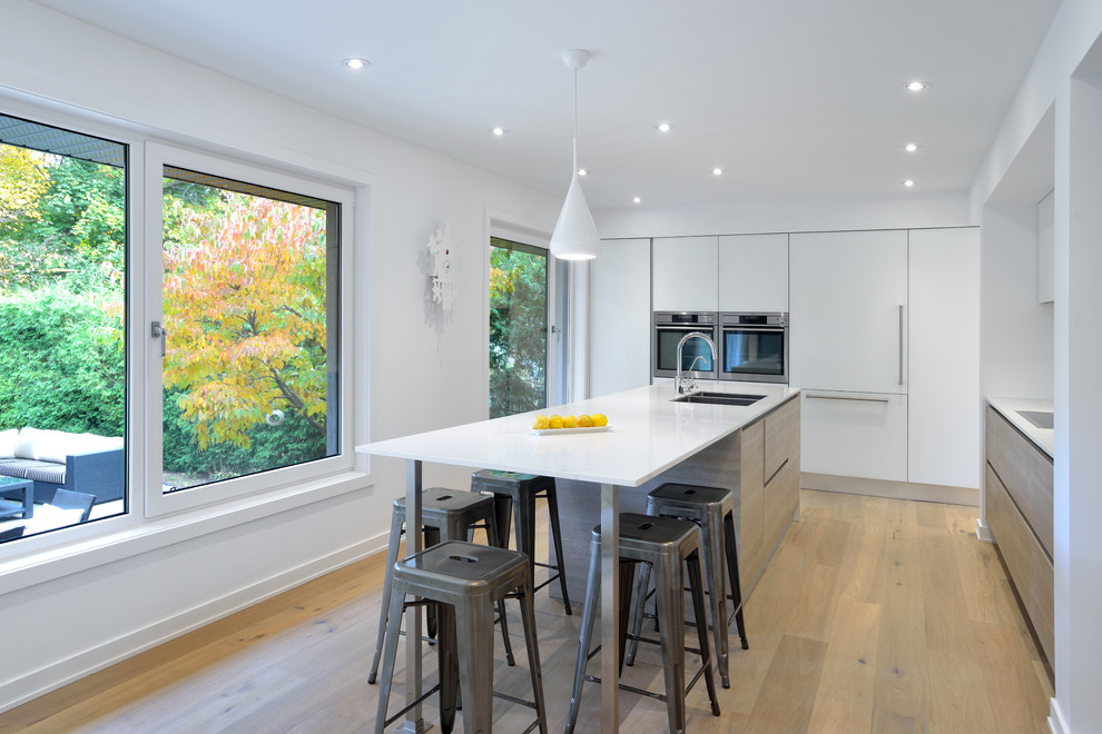 Photo of a contemporary l-shaped kitchen in Toronto with flat-panel cabinets, white cabinets, with island, a double-bowl sink, marble benchtops, white splashback, stone slab splashback, stainless steel appliances and light hardwood floors.