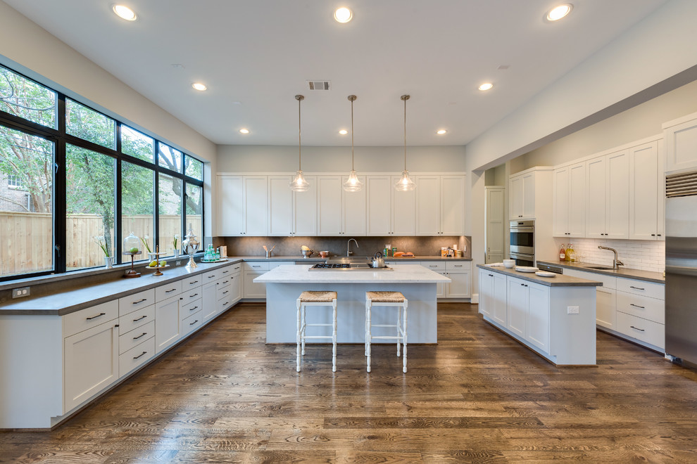 This is an example of a large transitional u-shaped open plan kitchen in Houston with a farmhouse sink, shaker cabinets, grey cabinets, limestone benchtops, grey splashback, stone tile splashback, stainless steel appliances, medium hardwood floors and multiple islands.