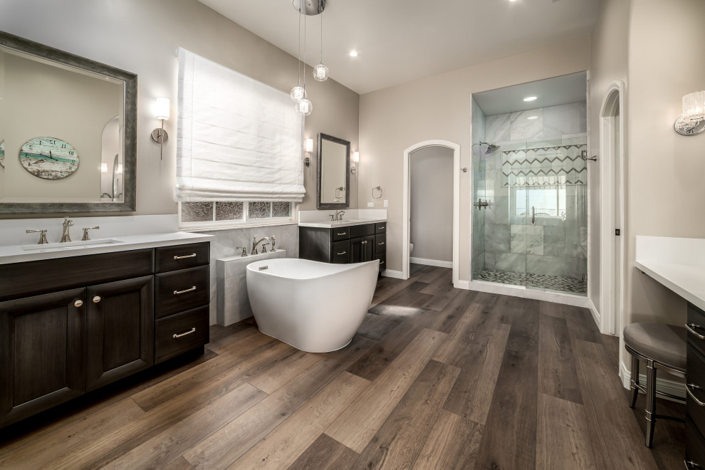 This is an example of a large transitional master bathroom in San Diego with dark wood cabinets, a freestanding tub, an open shower, white tile, marble, beige walls, an undermount sink, brown floor, a hinged shower door, white benchtops, a shower seat, a freestanding vanity, recessed-panel cabinets, a one-piece toilet, laminate floors, engineered quartz benchtops and a double vanity.