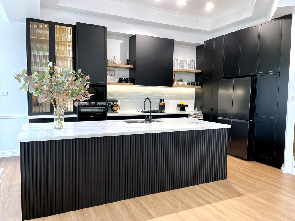 This is an example of a large modern l-shaped open plan kitchen in Sydney with an undermount sink, shaker cabinets, black cabinets, quartz benchtops, white splashback, subway tile splashback, black appliances, medium hardwood floors, with island, brown floor, white benchtop and recessed.