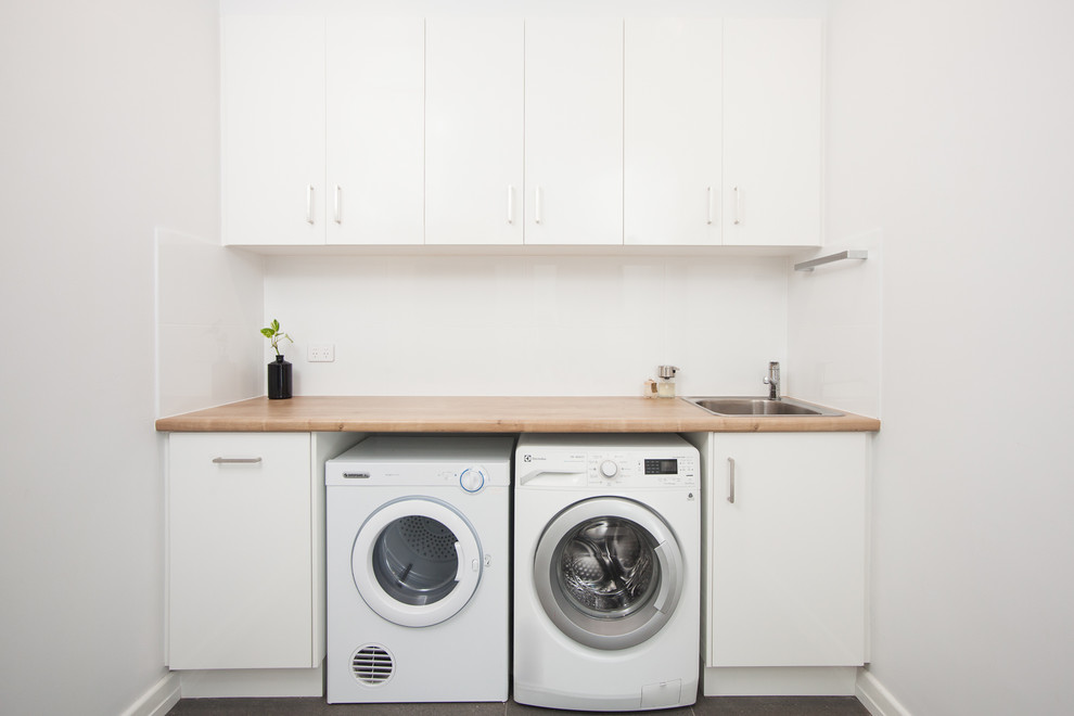 Modern single-wall dedicated laundry room in Brisbane with a drop-in sink, flat-panel cabinets, white cabinets, laminate benchtops, white walls, porcelain floors, a side-by-side washer and dryer, grey floor and brown benchtop.