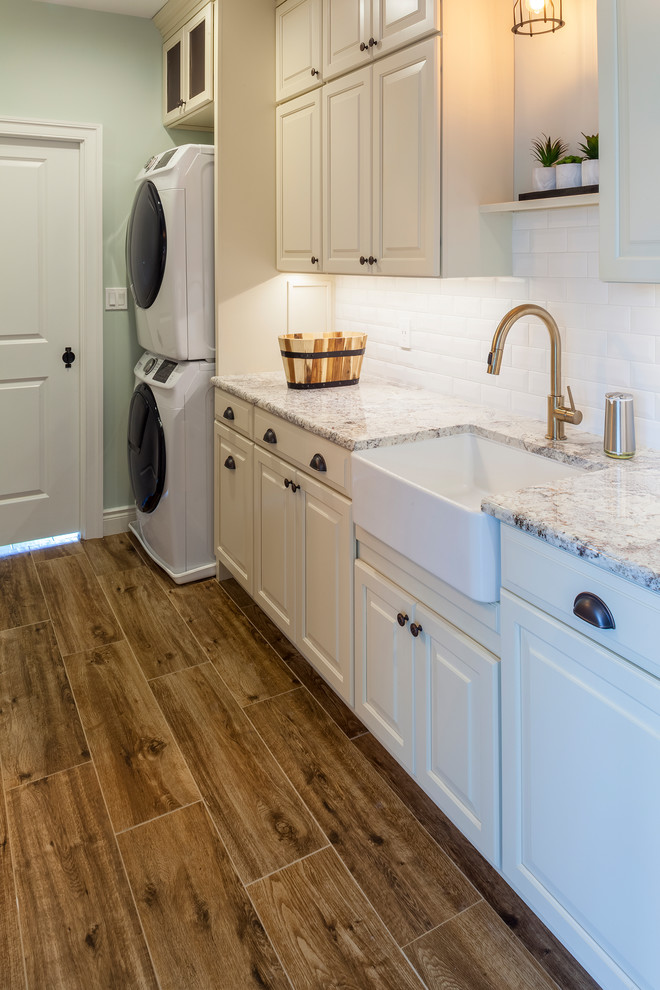 Photo of a mid-sized transitional single-wall utility room in Baltimore with a farmhouse sink, raised-panel cabinets, white cabinets, granite benchtops, grey walls, porcelain floors, a stacked washer and dryer, brown floor and grey benchtop.