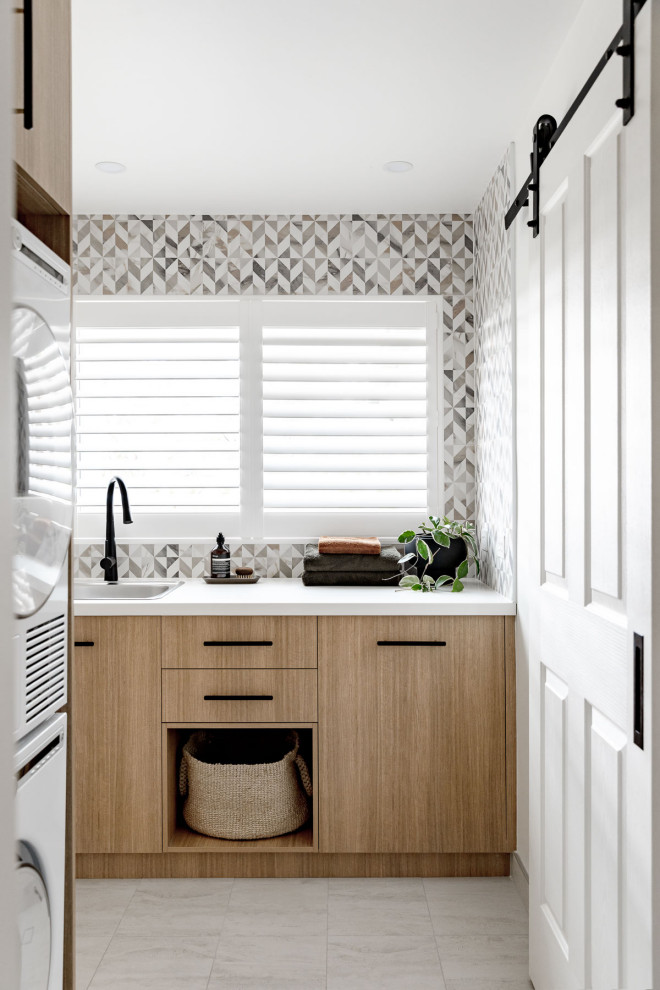 Example of a mid-sized trendy ceramic tile and white floor dedicated laundry room design in Sydney with a single-bowl sink, light wood cabinets, quartz countertops, porcelain backsplash, white walls, a stacked washer/dryer and white countertops