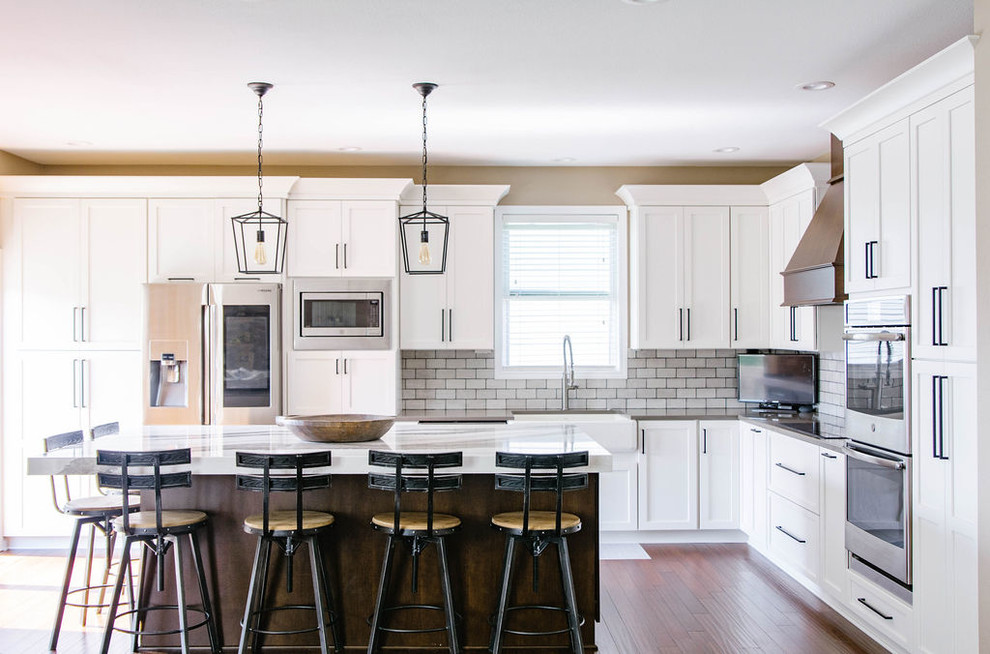 Kitchen - transitional l-shaped medium tone wood floor kitchen idea in Omaha with a farmhouse sink, shaker cabinets, white cabinets, gray backsplash, subway tile backsplash, stainless steel appliances, an island and gray countertops