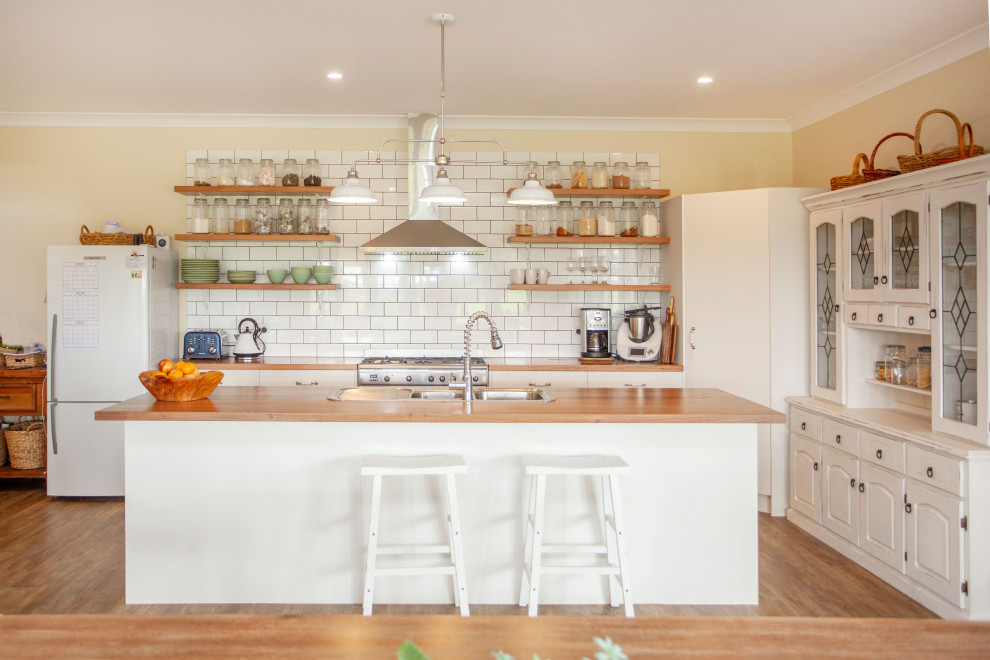 Photo of a country l-shaped kitchen in Other with a drop-in sink, flat-panel cabinets, white cabinets, wood benchtops, white splashback, subway tile splashback, stainless steel appliances, medium hardwood floors, with island, brown floor and brown benchtop.
