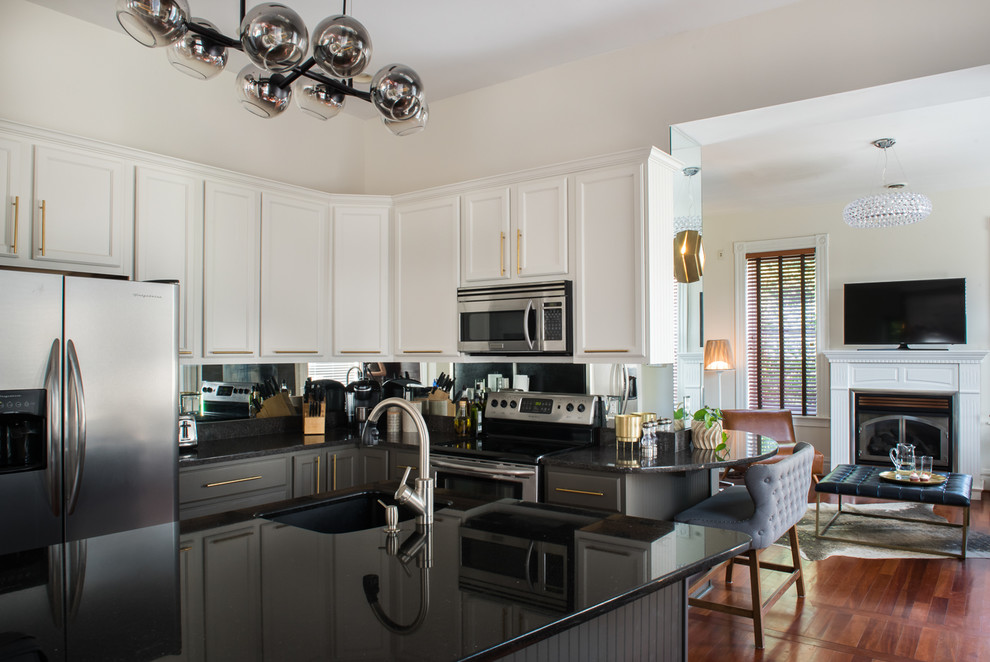 Inspiration for a mid-sized transitional l-shaped kitchen in St Louis with an undermount sink, flat-panel cabinets, granite benchtops, metallic splashback, mirror splashback, stainless steel appliances, medium hardwood floors, with island, brown floor, black benchtop and white cabinets.