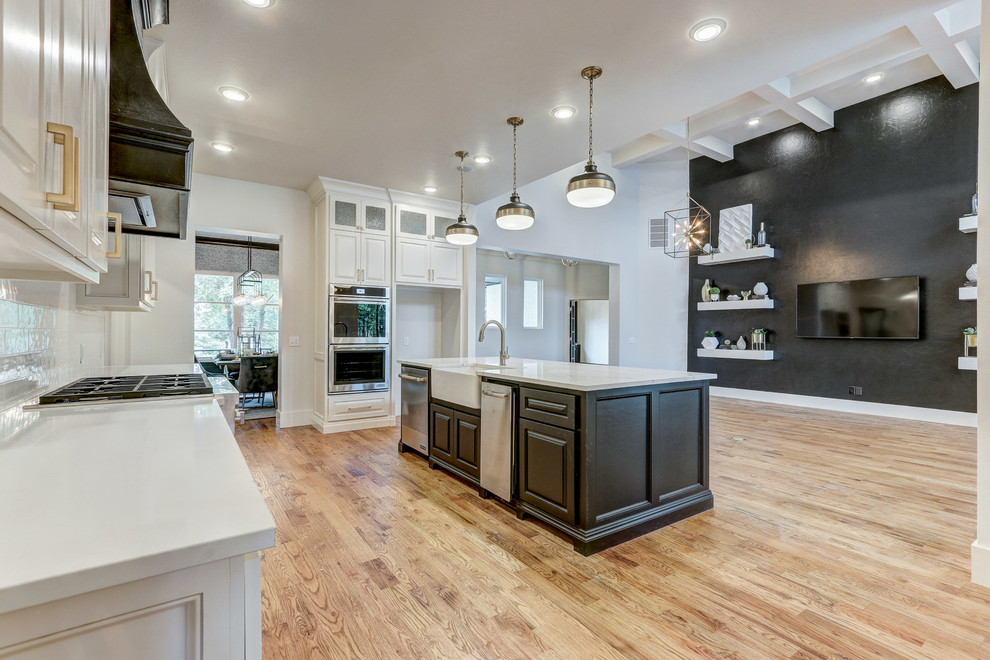 Inspiration for a large country l-shaped open plan kitchen in Oklahoma City with a belfast sink, flat-panel cabinets, black cabinets, quartz worktops, white splashback, metro tiled splashback, stainless steel appliances, medium hardwood flooring, an island, brown floors and white worktops.