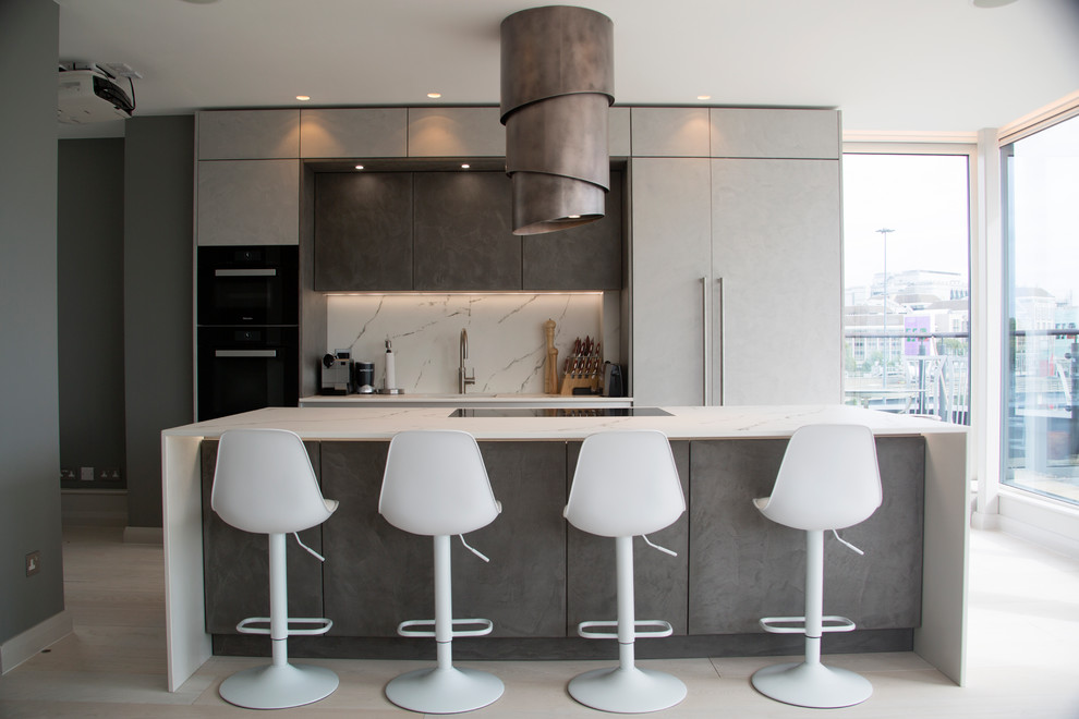 This is an example of a contemporary kitchen in Essex with flat-panel cabinets, grey cabinets, white splashback, black appliances, with island, grey floor and white benchtop.