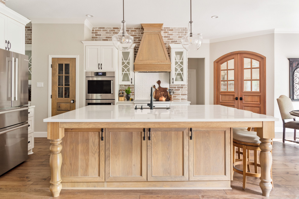 Photo of a large transitional l-shaped open plan kitchen in Dallas with a farmhouse sink, recessed-panel cabinets, white cabinets, quartz benchtops, red splashback, brick splashback, stainless steel appliances, medium hardwood floors, with island, brown floor and white benchtop.