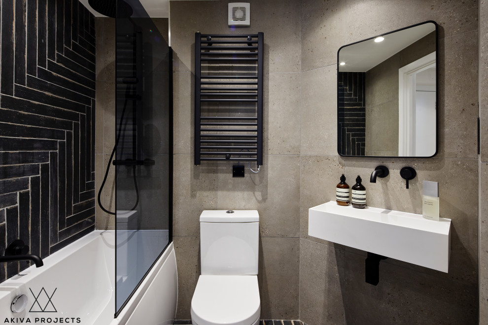 Photo of a small industrial kids bathroom in London with a drop-in tub, a shower/bathtub combo, a two-piece toilet, gray tile, porcelain tile, grey walls, porcelain floors, a wall-mount sink, black floor, a hinged shower door and a single vanity.