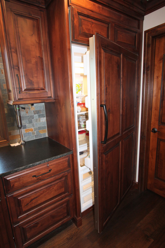 Large mountain style medium tone wood floor and brown floor kitchen photo with raised-panel cabinets, medium tone wood cabinets, granite countertops, multicolored backsplash, stone tile backsplash, paneled appliances, an island and black countertops