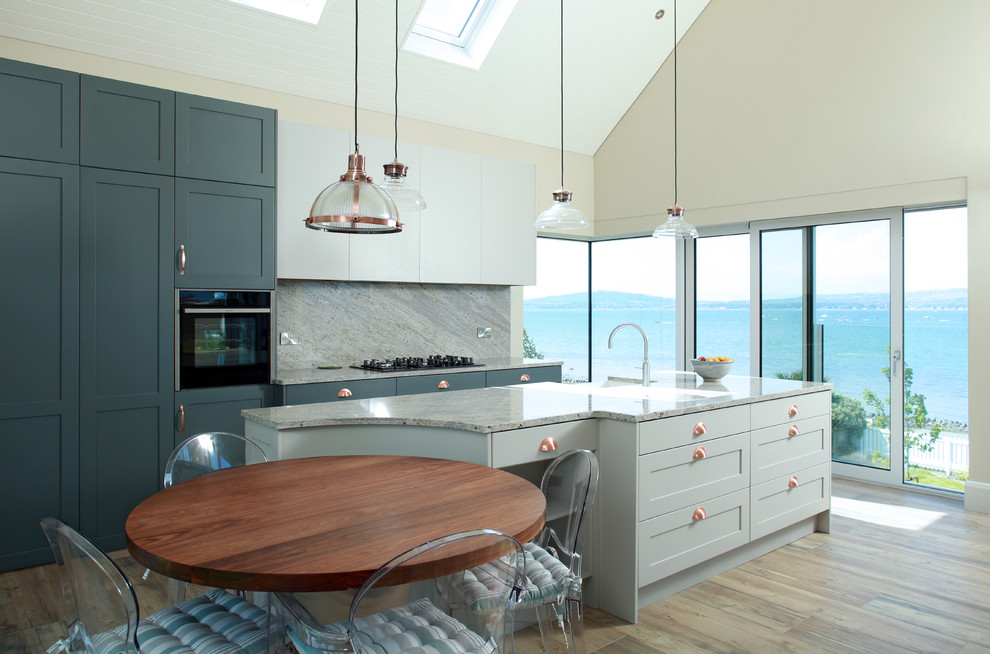 This is an example of a mid-sized beach style kitchen in London with shaker cabinets, marble benchtops, grey splashback, marble splashback, stainless steel appliances, with island, grey benchtop, a farmhouse sink, blue cabinets, light hardwood floors and brown floor.