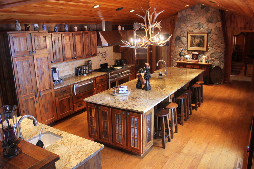 This is an example of a large country single-wall eat-in kitchen in Denver with a farmhouse sink, recessed-panel cabinets, distressed cabinets, granite benchtops, multi-coloured splashback, stone tile splashback, stainless steel appliances, light hardwood floors and multiple islands.