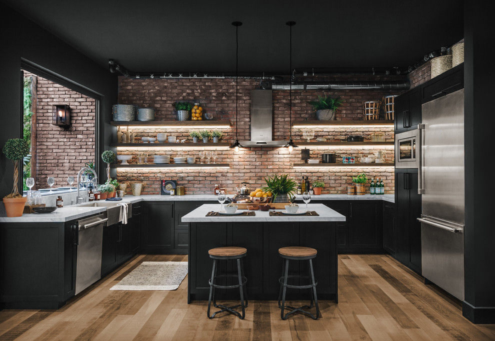 This is an example of a large industrial u-shaped kitchen in Los Angeles with laminate floors, a farmhouse sink, shaker cabinets, black cabinets, red splashback, brick splashback, stainless steel appliances, with island, brown floor and white benchtop.