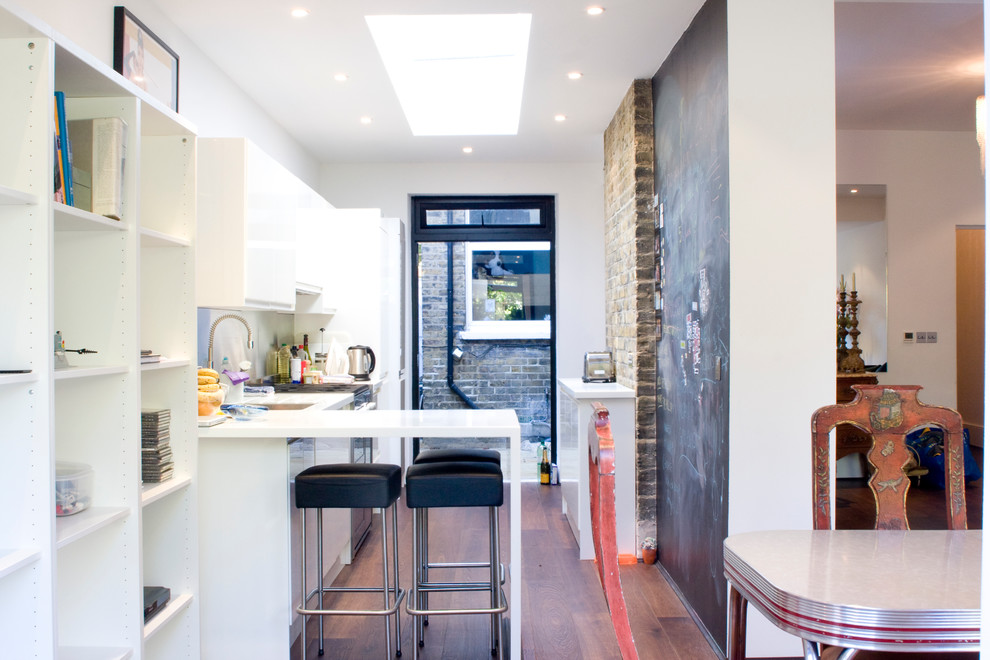 Mid-sized contemporary single-wall eat-in kitchen in London with an integrated sink, flat-panel cabinets, white cabinets, white splashback, glass tile splashback, dark hardwood floors, a peninsula, solid surface benchtops, panelled appliances and white benchtop.