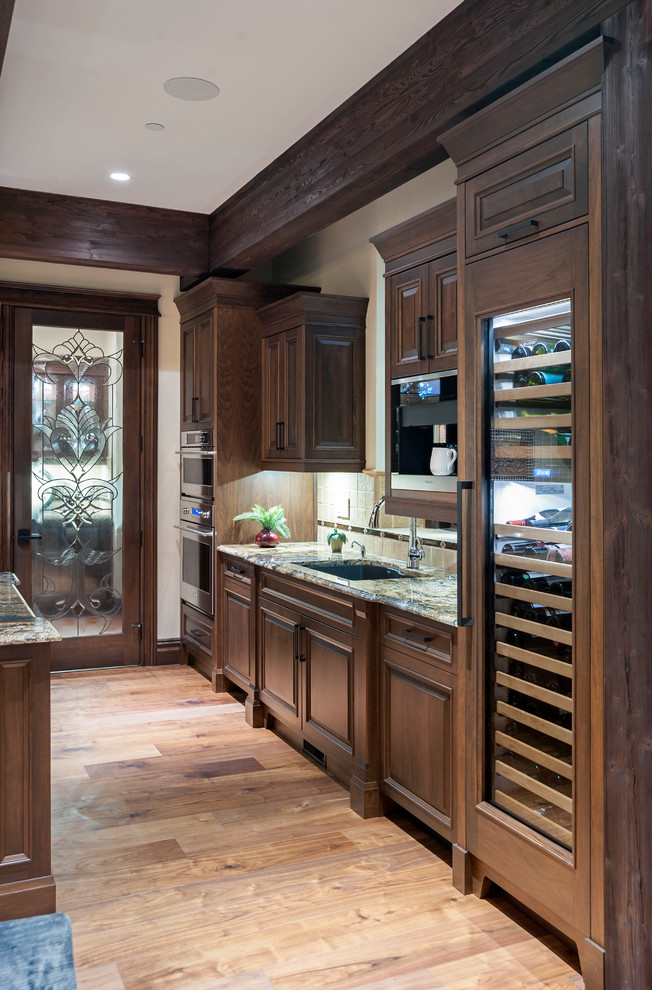 Inspiration for a large rustic l-shaped medium tone wood floor and brown floor open concept kitchen remodel in Calgary with brown cabinets, an undermount sink, raised-panel cabinets, granite countertops, beige backsplash, stone tile backsplash, paneled appliances and an island