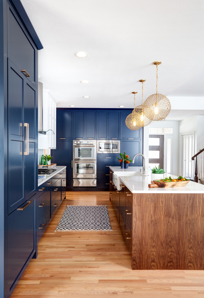 Photo of a large transitional galley eat-in kitchen in Minneapolis with a farmhouse sink, recessed-panel cabinets, blue cabinets, quartz benchtops, white splashback, porcelain splashback, stainless steel appliances, medium hardwood floors, with island and white benchtop.