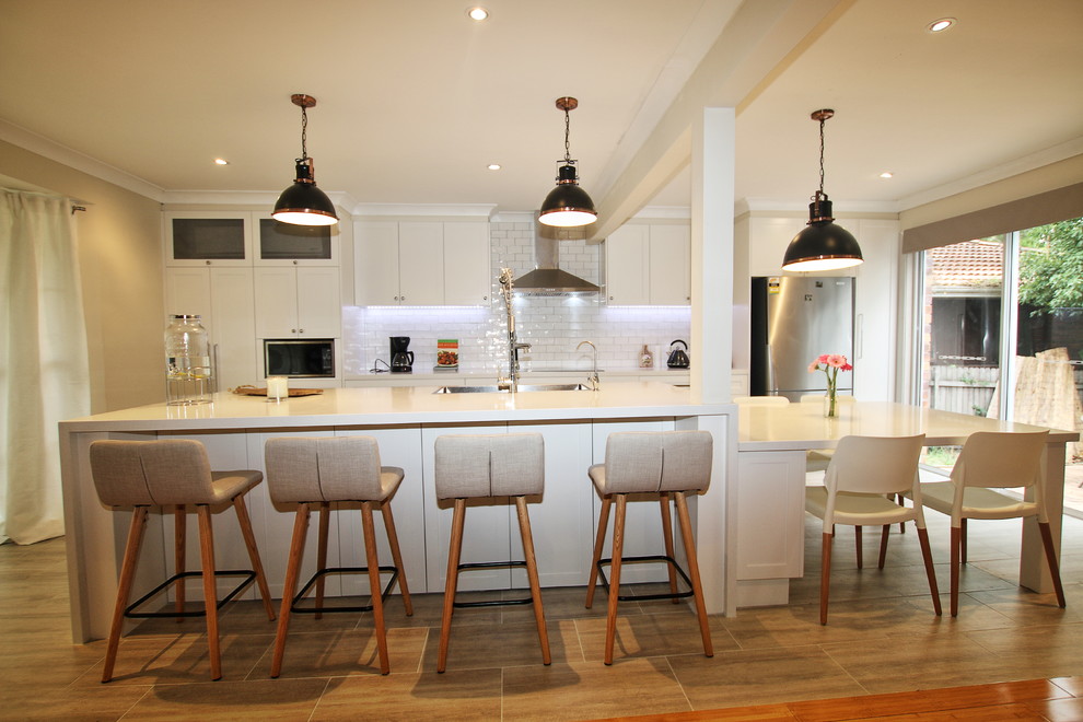 This is an example of a large transitional single-wall kitchen in Sydney with a double-bowl sink, shaker cabinets, white cabinets, quartz benchtops, white splashback, subway tile splashback, stainless steel appliances, porcelain floors and with island.