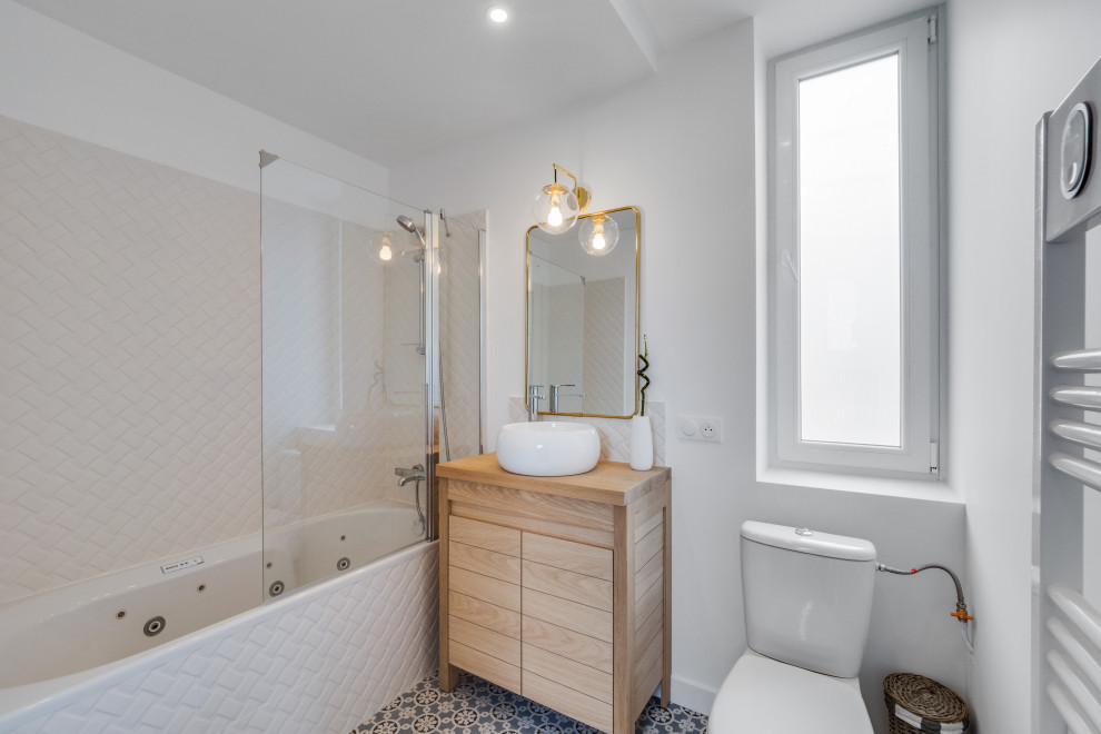 Mid-sized traditional master bathroom in Paris with flat-panel cabinets, light wood cabinets, an undermount tub, a shower/bathtub combo, a one-piece toilet, white tile, ceramic tile, white walls, cement tiles, a drop-in sink, wood benchtops, blue floor, a hinged shower door, beige benchtops, a single vanity and a freestanding vanity.