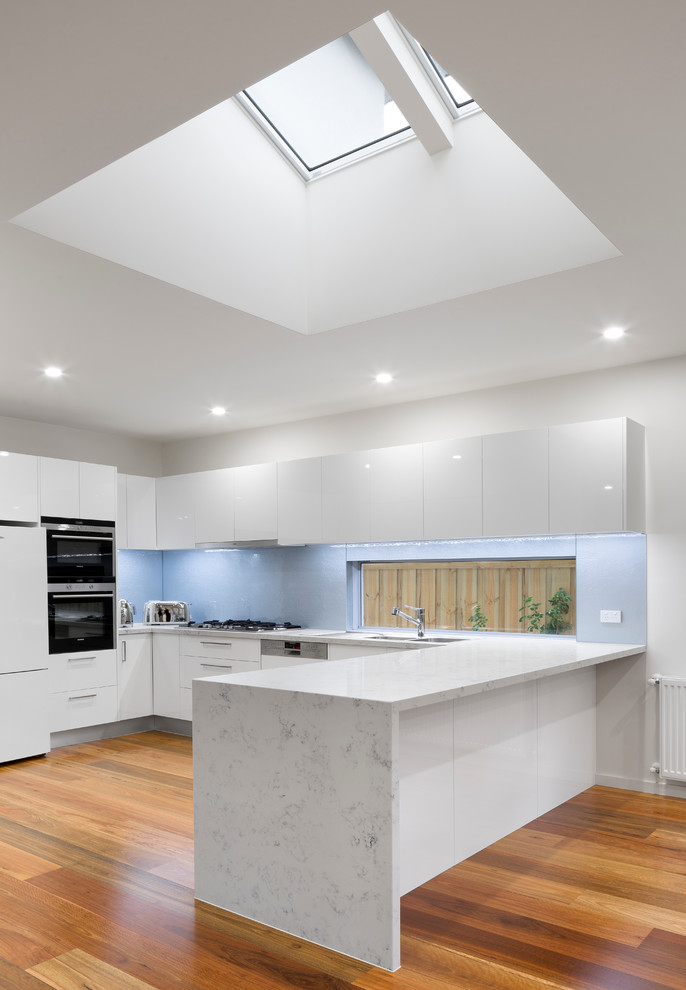 Photo of a mid-sized modern u-shaped open plan kitchen in Melbourne with a double-bowl sink, flat-panel cabinets, white cabinets, marble benchtops, white splashback, metal splashback, white appliances, light hardwood floors and no island.