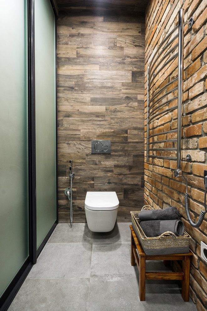 Photo of a mid-sized industrial powder room in Other with glass-front cabinets, dark wood cabinets, a wall-mount toilet, brown tile, porcelain tile, grey walls, porcelain floors, a drop-in sink, tile benchtops and grey floor.