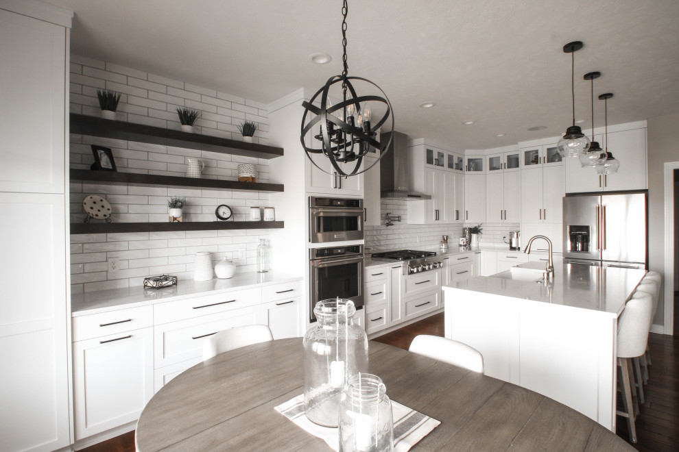 Transitional u-shaped kitchen photo with a farmhouse sink, shaker cabinets, white cabinets, quartz countertops, white backsplash, porcelain backsplash, stainless steel appliances, an island and white countertops