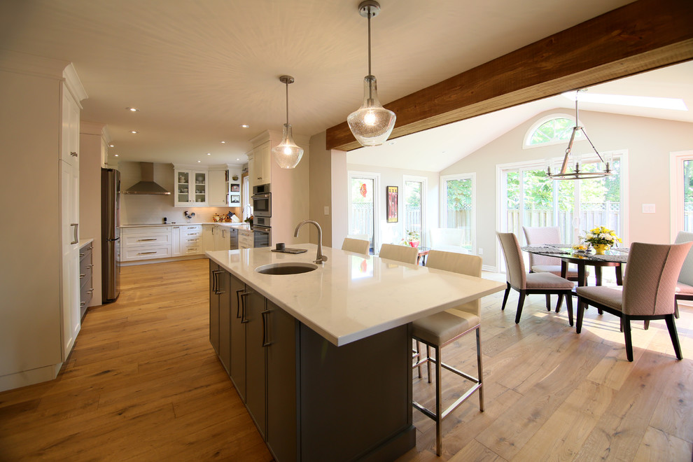 Example of a large transitional l-shaped medium tone wood floor and brown floor eat-in kitchen design with shaker cabinets, white cabinets, an island, an undermount sink, stainless steel appliances, solid surface countertops and white backsplash