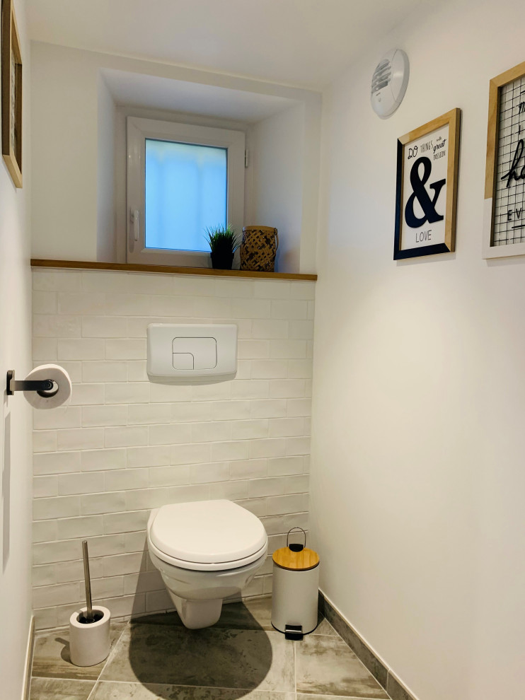 This is an example of a mid-sized country powder room in Paris with a wall-mount toilet, white tile, ceramic tile, white walls, ceramic floors and grey floor.