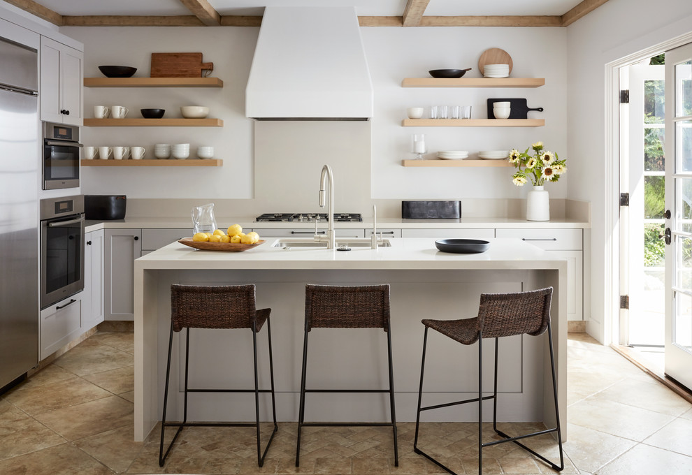 Design ideas for a mid-sized beach style l-shaped kitchen in San Luis Obispo with a double-bowl sink, stainless steel appliances, with island, beige floor, open cabinets, light wood cabinets, white splashback, travertine floors and white benchtop.