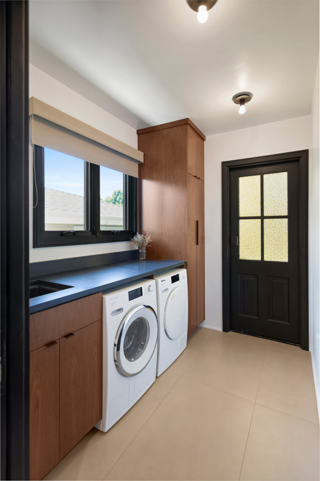 Design ideas for a small contemporary utility room in Los Angeles with a submerged sink, flat-panel cabinets, medium wood cabinets, granite worktops, white walls, porcelain flooring, a side by side washer and dryer, grey floors and black worktops.