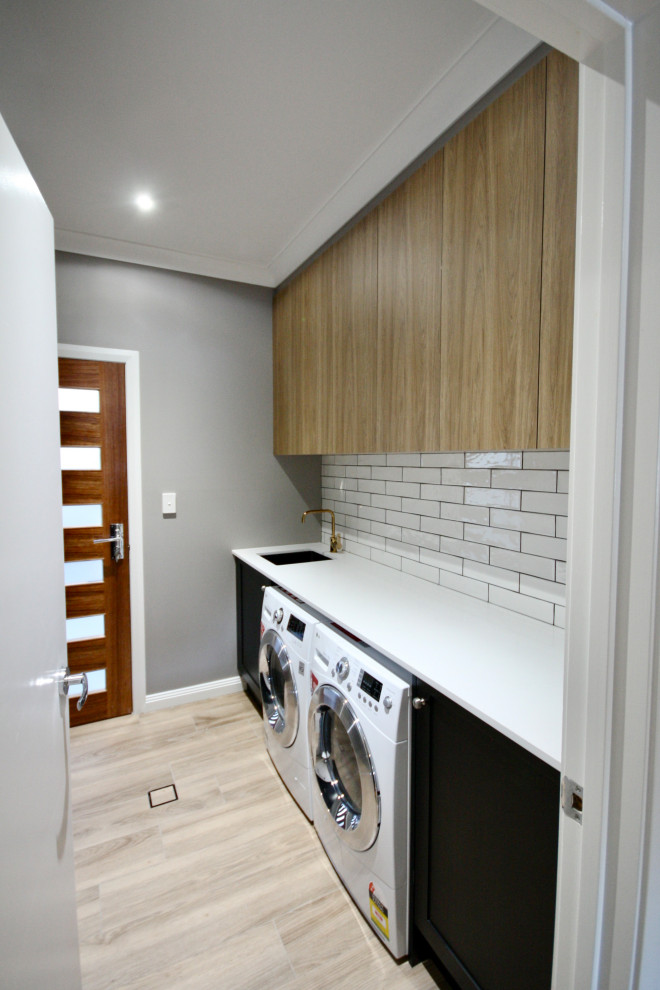 This is an example of a mid-sized transitional galley dedicated laundry room in Sydney with an undermount sink, shaker cabinets, black cabinets, quartz benchtops, white splashback, subway tile splashback, grey walls, light hardwood floors, a side-by-side washer and dryer, brown floor, white benchtop and recessed.