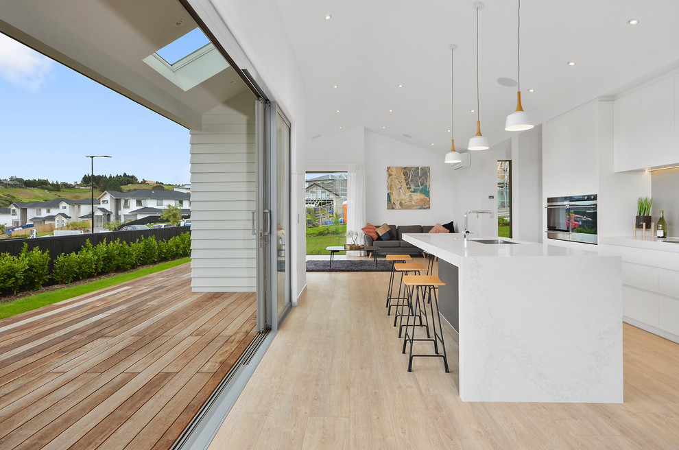 Design ideas for a large modern single-wall eat-in kitchen in Auckland with a drop-in sink, raised-panel cabinets, white cabinets, grey splashback, glass sheet splashback, stainless steel appliances, light hardwood floors, with island, brown floor and white benchtop.